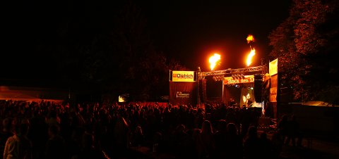 Die Bühne auf dem Rock den Deich Festival in der Nacht. Darauf ein singender Künstler vor dem Publikum. Über der Bühne sind Flammen.